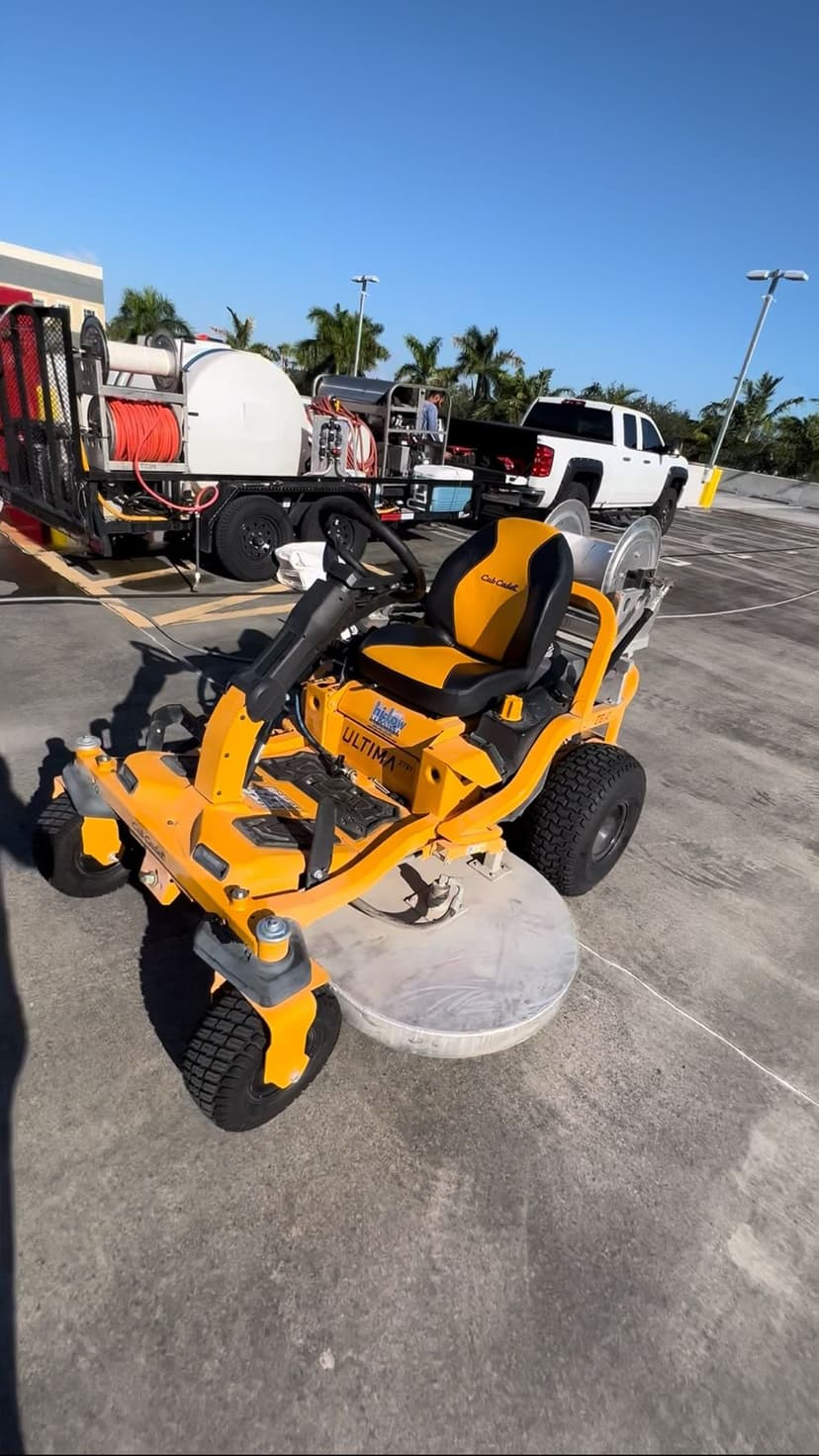 Yellow ride-on lawn mower parked on concrete with landscaping equipment in the background.